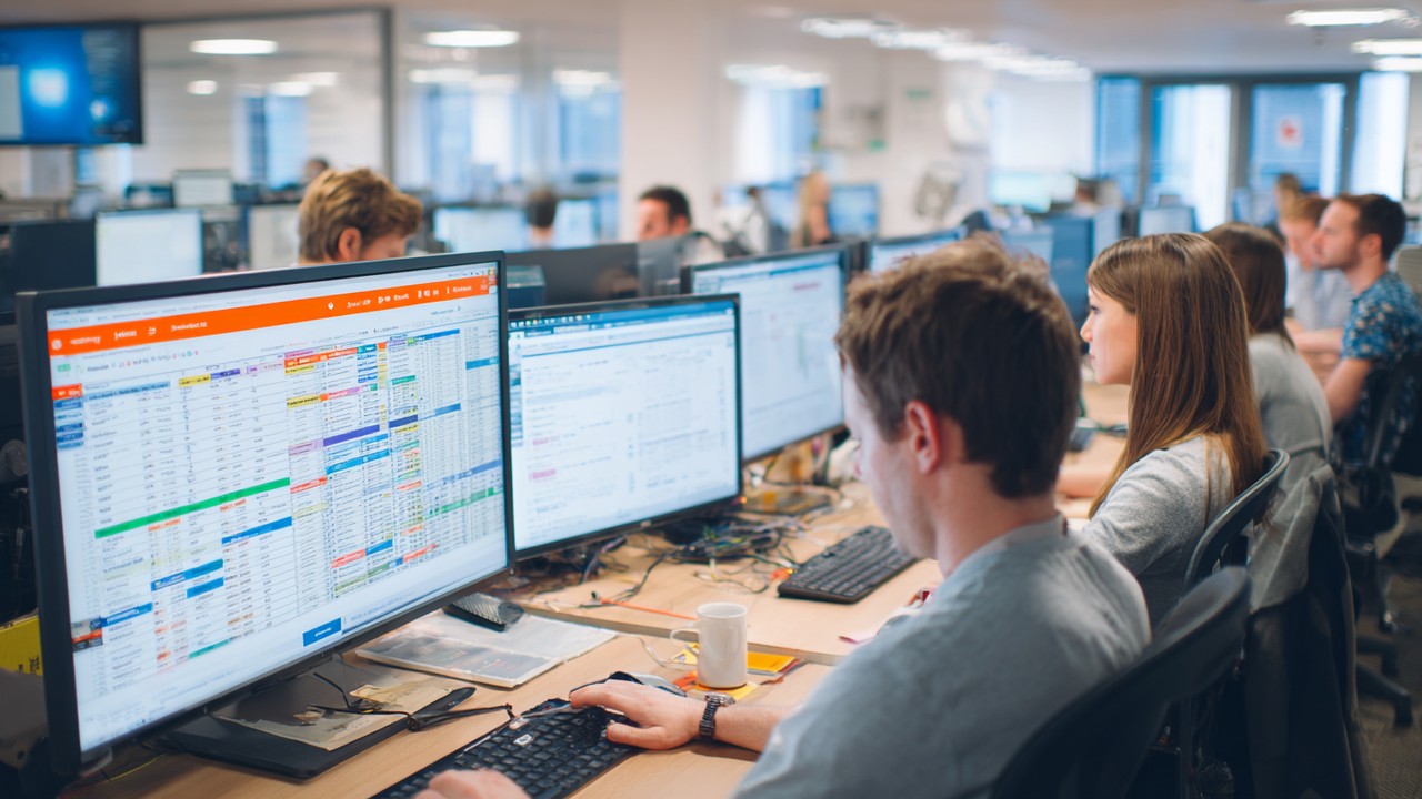 Busy office scene with SEO team members working on computers displaying spreadsheets and SEO management tools, highlighting enterprise SEO challenges.