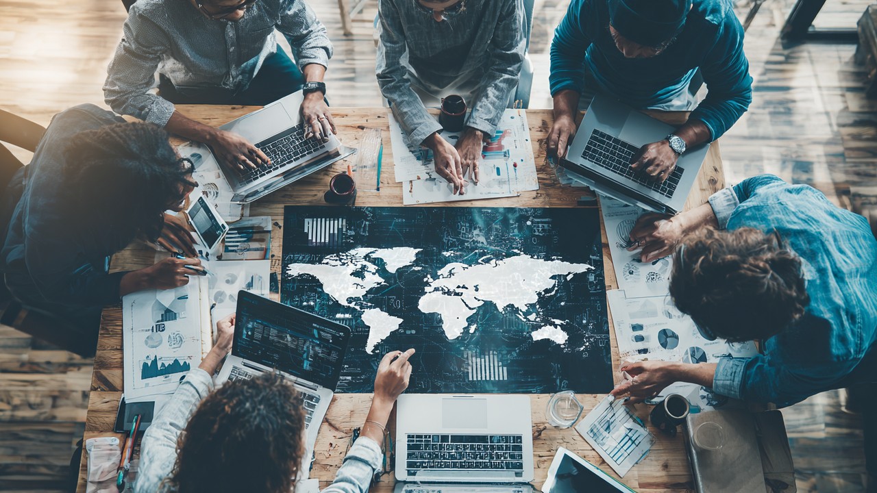 Diverse digital marketers collaborating around a table with laptops, analyzing global data charts and maps in a modern office.