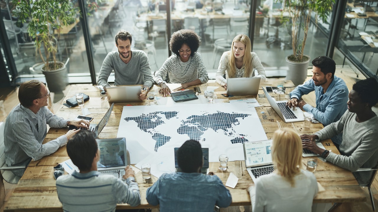Diverse marketing team collaborating around a table with laptops and digital devices, analyzing global SEO strategy charts and world maps in a modern office.