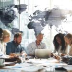 Diverse professionals collaborating around a modern office table, analyzing global data and digital marketing charts with laptops and tablets.