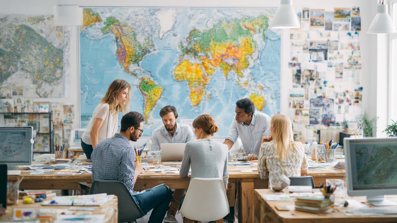 Travel agency team reviewing localized travel content on screens with destination visuals, world maps, and travel memorabilia in a bright office.